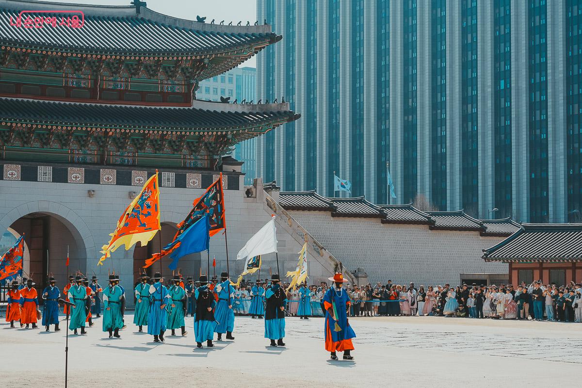 회색빛 고층 빌딩 앞을 지나가는, 푸른색과 붉은색 전통 의상을 입고 깃발을 든 수문군들의 횡대 사진