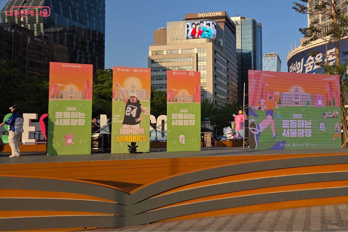 서울광장 메인무대 위에 &lsquo;2026 운동하는 서울광장&rsquo;과 &lsquo;에어로빅 AEROBICS&rsquo; 안내 패널이 세워져 있다. 무대 주변에는 대형 배경판과 행사 장식물이 설치되어 있고, 강사가 프로그램 시작 전 무대에서 준비하고 있다.