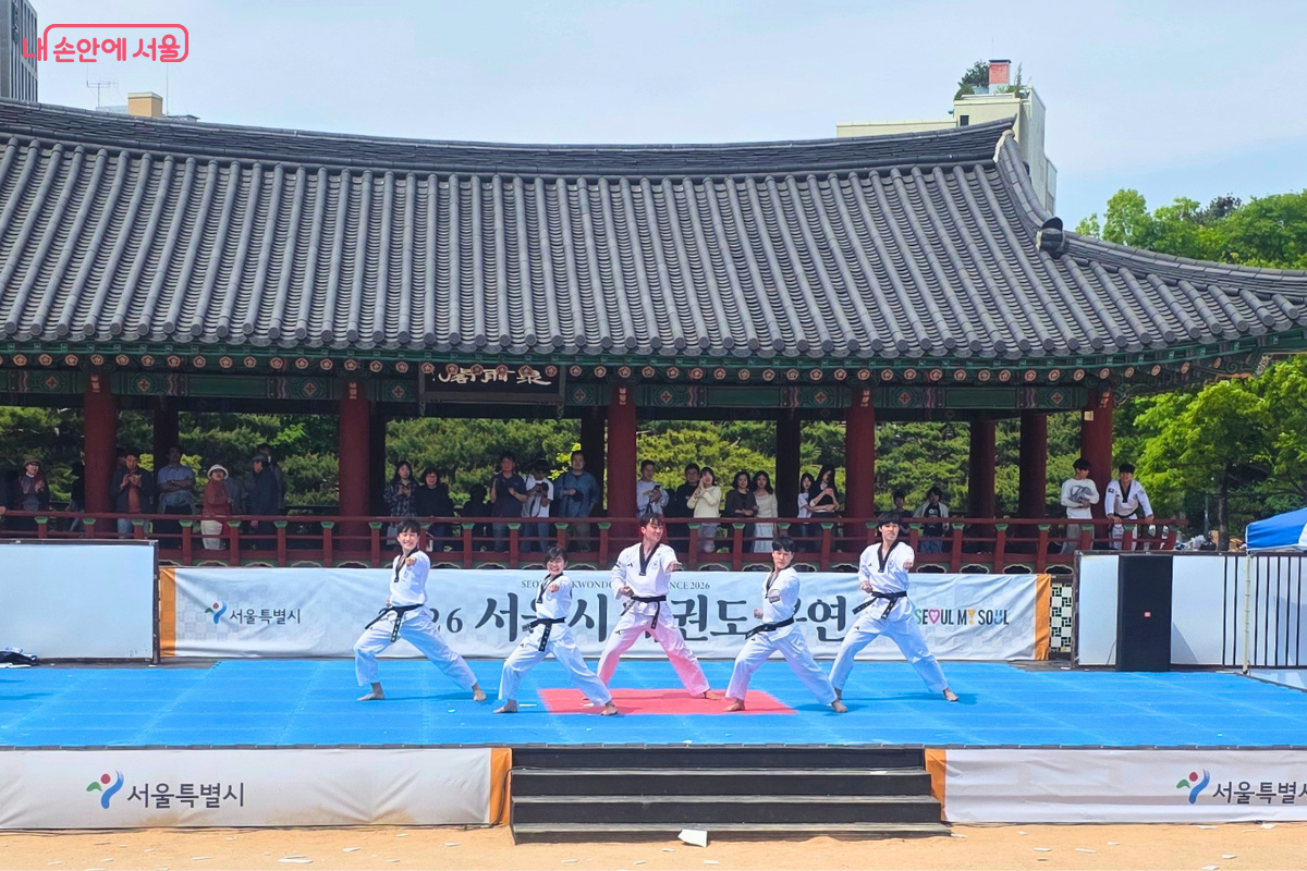 남산골한옥마을 천우각 무대에서 흰색 도복에 검은 띠를 맨 태권도 시범단원 5명이 일렬로 서서 품새 동작을 선보이고 있다. 전통 한옥 지붕을 배경으로 파란 매트 위에서 공연 중이며, 무대 앞에는 '서울특별시' 로고가 붙은 흰색 가림막이 있다. 배경의 정자 뒤편에서는 관람객들이 공연을 지켜보고 있다.