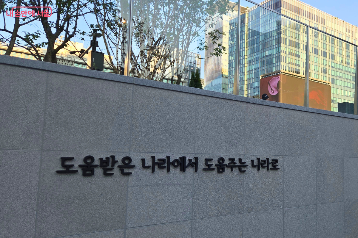 프리덤 홀로 내려가는 회색 벽면에 &lsquo;도움받은 나라에서 도움주는 나라로&rsquo;라는 문구가 검은 글씨로 설치되어 있고, 위쪽으로 광화문 일대의 건물과 나무가 보인다.