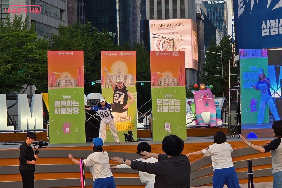 &lsquo;2026 운동하는 서울광장&rsquo; 무대 위에서 에어로빅 강사가 팔을 뻗으며 동작을 시범 보이고 있다. 무대 앞 시민들은 강사의 움직임을 따라 하며 운동에 참여하고, 오른쪽 대형 화면에는 강사의 모습이 확대되어 보인다.
