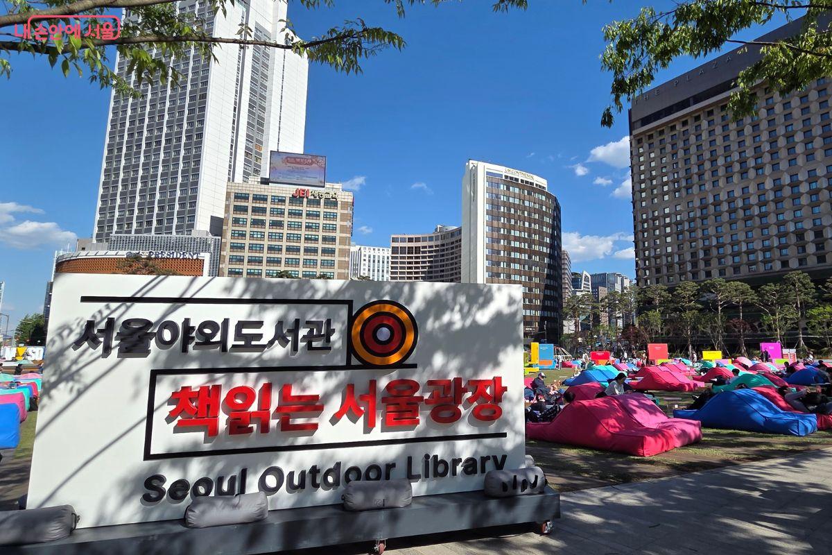 서울광장 앞에 &lsquo;서울야외도서관 책읽는 서울광장&rsquo;이라고 적힌 대형 안내 사인이 설치되어 있고, 뒤편 잔디광장에는 분홍색과 파란색 빈백에 앉아 책을 읽는 시민들이 보인다. 배경으로 서울 도심의 고층 건물과 푸른 하늘이 펼쳐져 있다.