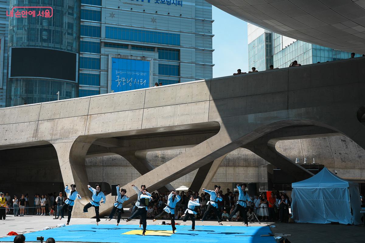 DDP 광장 야외 무대에서 하늘색 도복의 퍼포머들이 동시에 점프하는 장면, 뒤편으로 관람객과 도심 빌딩