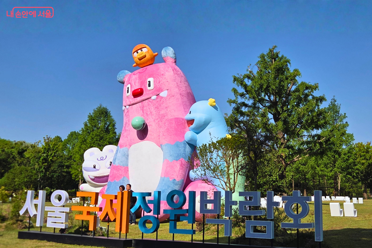 맑고 파란 하늘 아래 넓은 잔디 광장을 배경으로 거대한 서울시 캐릭터 해치와 소울프렌즈 조형물이 서 있다. 조형물 앞에는 '서울국제정원박람회'라는 글자가 입체적으로 세워져 있다.