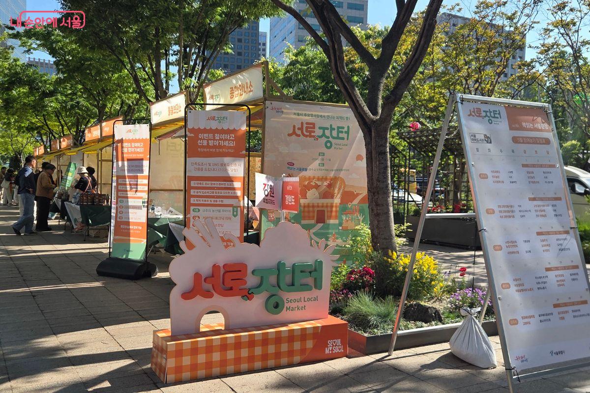 서울광장 한켠에 &lsquo;서로장터&rsquo;라고 적힌 주황색 안내 조형물과 배너가 설치되어 있고, 뒤편으로 지역 상생 장터 부스들이 줄지어 있다. 시민들이 부스를 둘러보며 행사장을 오가고 있다.