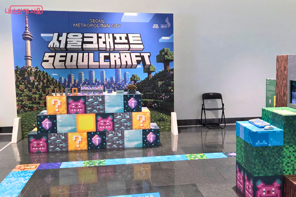 서울갤러리 내 '서울크래프트(SEOULCRAFT)' 체험 전시 공간이 보인다. 배경에는 남산타워와 서울 도심이 그려진 'SEOUL METROPOLITAN CITY 서울크래프트 SEOULCRAFT'라고 적힌 대형 배너가 걸려 있다. 전면에는 물음표와 해치 캐릭터가 그려진 픽셀 블록 형태의 큐브들이 쌓여 있고, 바닥에는 블록 패턴의 매트가 깔려 있다. 우측에는 키보드와 마우스가 올려진 블록형 테이블이 보인다.