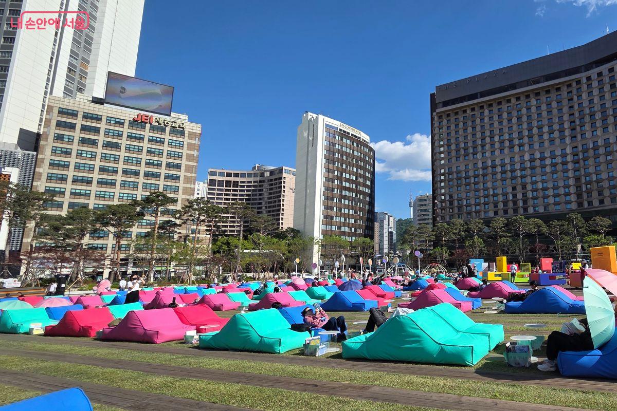 서울광장 잔디밭 전체에 분홍색, 파란색, 청록색 빈백이 넓게 배치되어 있고, 시민들이 빈백에 기대어 책을 읽거나 휴식을 취하고 있다. 배경으로 서울 도심의 호텔과 고층 건물, 맑은 하늘이 보인다.