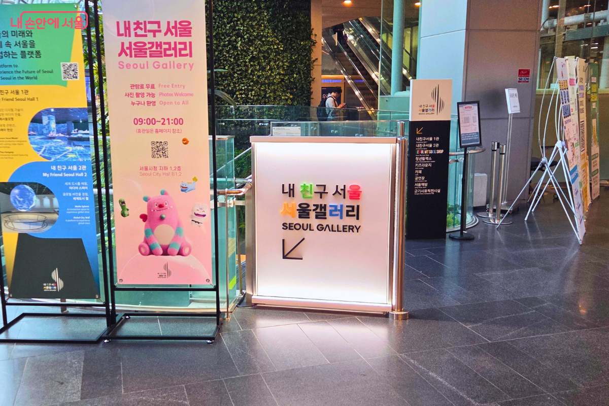서울시청 1층에 설치된 '내 친구 서울 서울갤러리' 입구 안내판이 보인다. 분홍색 배경의 해치 캐릭터가 그려진 배너에는 '관람료 무료', '사진 촬영 가능', '누구나 환영'이라는 문구와 운영 시간 '09:00~21:00'이 표시되어 있다. 배너 옆에는 흰색 안내판에 '내 친구 서울 서울갤러리 SEOUL GALLERY'라고 적혀 있고 화살표가 지하 방향을 가리키고 있다. 배경에는 에스컬레이터와 녹색 식물 벽이 보인다.