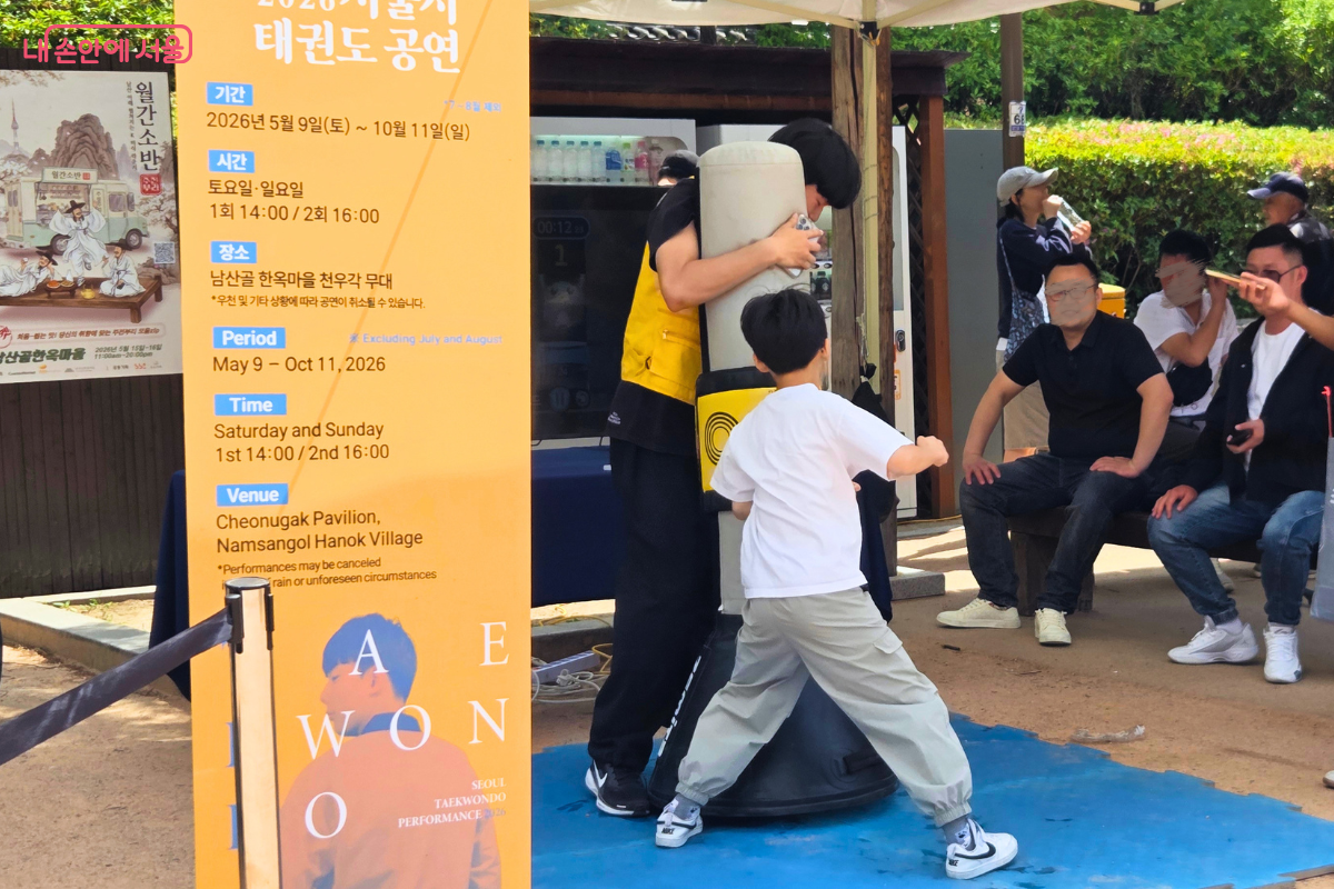 남산골한옥마을 태권도 체험존에서 어린이가 전자 타격 측정 기구를 이용해 발차기 체험을 하고 있다. 노란색 조끼를 입은 관계자가 에어백을 잡고 있으며, 흰색 티셔츠를 입은 어린이가 태권도 동작을 취하고 있다. 옆에는 '2026 서울시 태권도 공연'이라고 적힌 노란색 안내 배너가 세워져 있다.