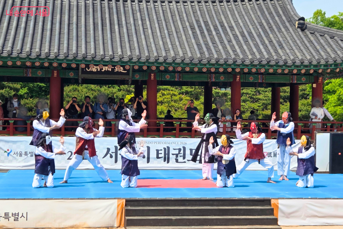 남산골한옥마을 천우각 무대에서 탈을 쓴 태권도 시범단이 품새 공연을 펼치고 있다. 하회탈 등 전통 탈을 쓴 채 흰색 바지에 색동 조끼를 입은 단원들이 군무를 선보이고 있으며, 배경에는 사진을 찍는 관람객들이 보인다. 무대 배너에는 '2026 서울시 태권도 공연'이라고 적혀 있다.