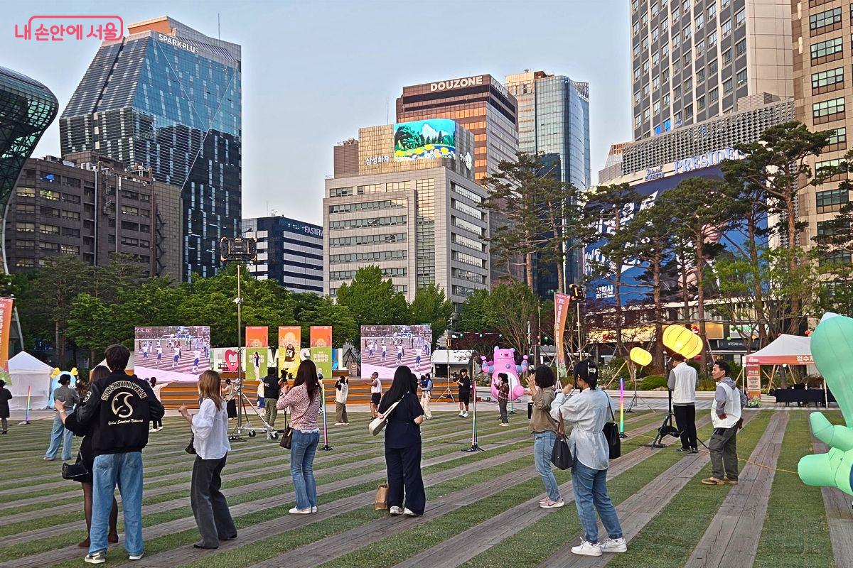 서울광장에 마련된 야외 무대 앞에서 시민들과 관광객들이 서서 에어로빅 프로그램에 참여하고 있다. 주변에는 고층 빌딩과 대형 전광판, 행사 부스와 조형물이 보이며, 퇴근길 도심 한복판에서 시민들이 함께 운동하는 모습이 펼쳐져 있다.