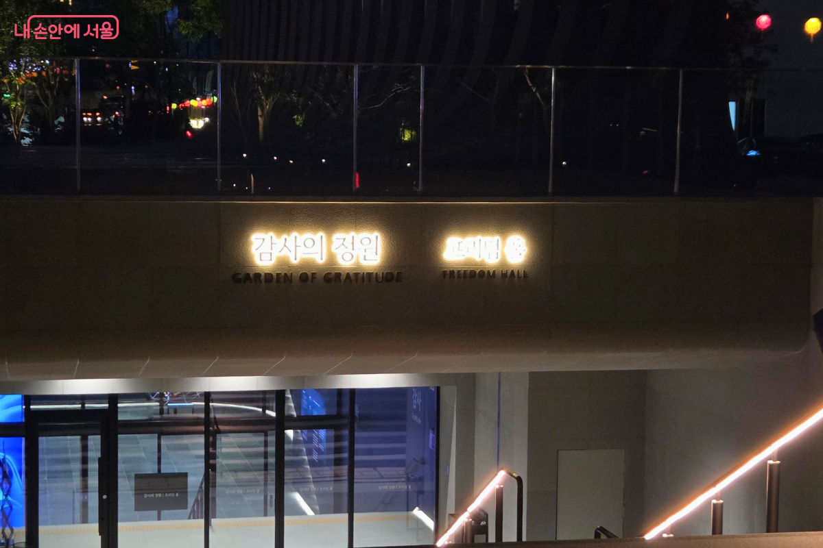 어두운 저녁 프리덤 홀 입구에서 '감사의 정원 프리덤 홀' 이름이 밝게 빛나고 있으며, 아래쪽 유리문 너머로 지하 공간이 보인다.