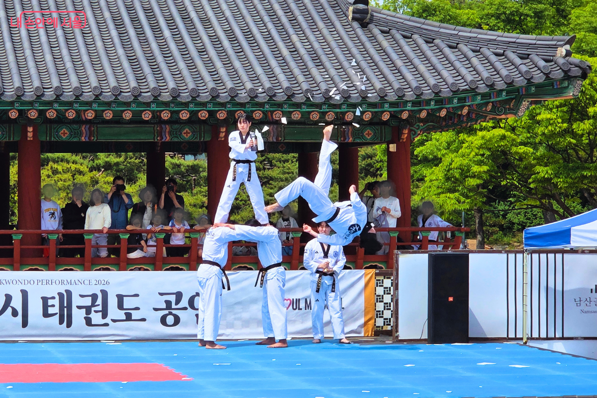 남산골한옥마을 천우각 무대에서 경민대학교 태권도 시범단이 고난도 격파 기술을 선보이고 있다. 한 단원이 다른 단원들이 만든 인간 탑 위에서 송판을 들고 있고, 다른 단원이 공중에서 뒤로 젖힌 채 발로 송판을 격파하고 있다. 격파된 송판 조각이 공중으로 흩날리고 있으며, 배경에는 관람객들이 보인다.