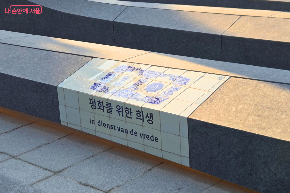 회색 석재 계단 사이에 흰색과 파란색 타일 장식이 설치되어 있고, 전면에는 &lsquo;평화를 위한 희생&rsquo;이라는 문구와 네덜란드어 문장이 함께 적혀 있다.