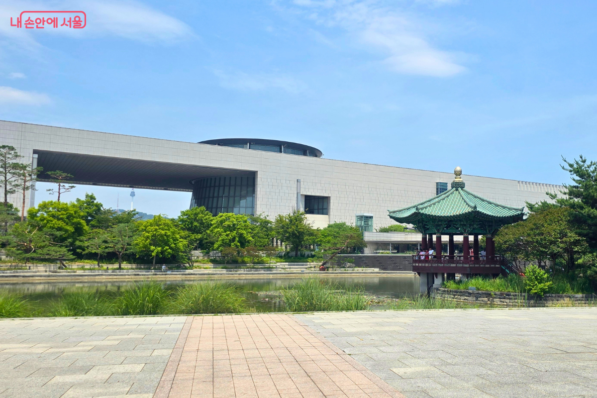국립중앙박물관 야외 연못과 정자, 뒤편의 현대적인 박물관 건물이 함께 보인다. 푸른 하늘 아래 연못 주변으로 나무와 수풀이 우거져 있다.
