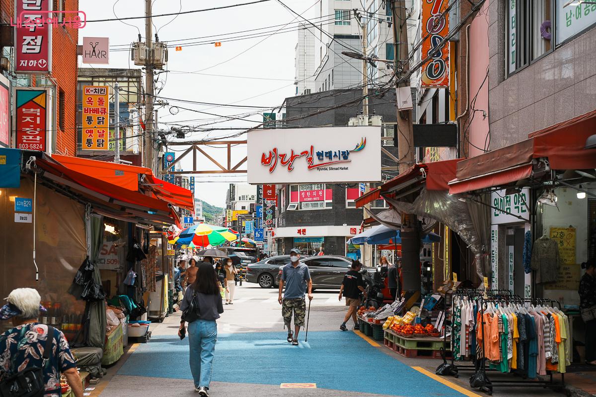 비단길현대시장 간판, 파란 바닥 골목길, 양쪽 상점들, 오가는 시민들