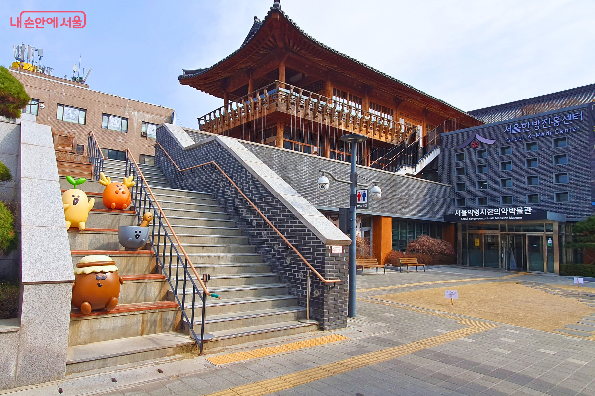 서울한방진흥센터 입구 전경이다. 계단 위로 전통 한옥 건물이 보이고, 오른쪽에는 서울약령시한의약박물관 출입구와 센터 건물 외벽 간판이 보인다. 계단 옆에는 귀여운 한방 캐릭터 조형물이 놓여 있다.