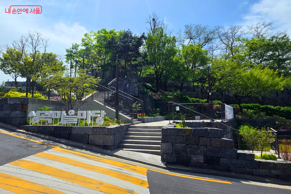 다산성곽길 안쪽의 성곽마을마당 입구가 보인다. 돌계단과 석축, 나무들이 어우러져 있고, 왼쪽에는 ‘성곽마을마당’ 글자 조형물이 설치되어 있다.