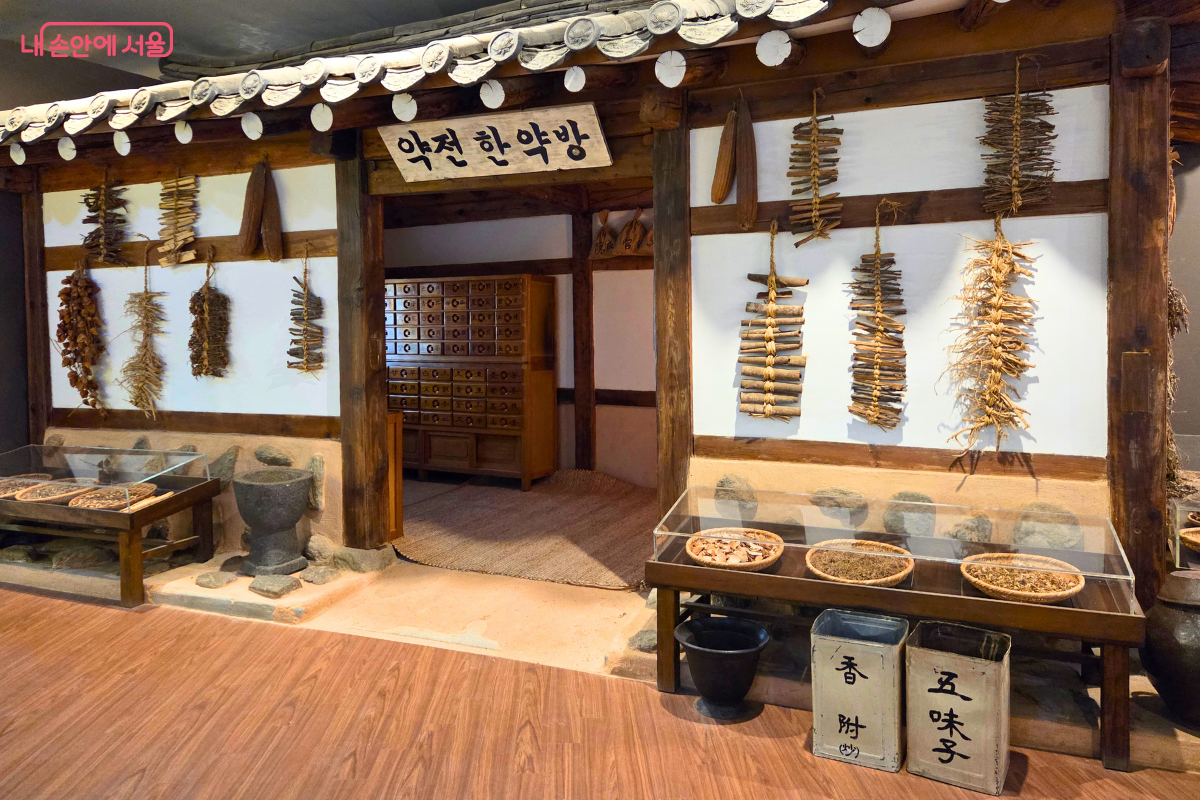 서울약령시한의약박물관 내부의 ‘약진 한약방’ 재현 전시 공간이다. 한옥 형태의 전시 구조물과 약재 서랍장, 절구, 말린 약재, 진열된 한약재가 전통 한약방 분위기를 보여준다.