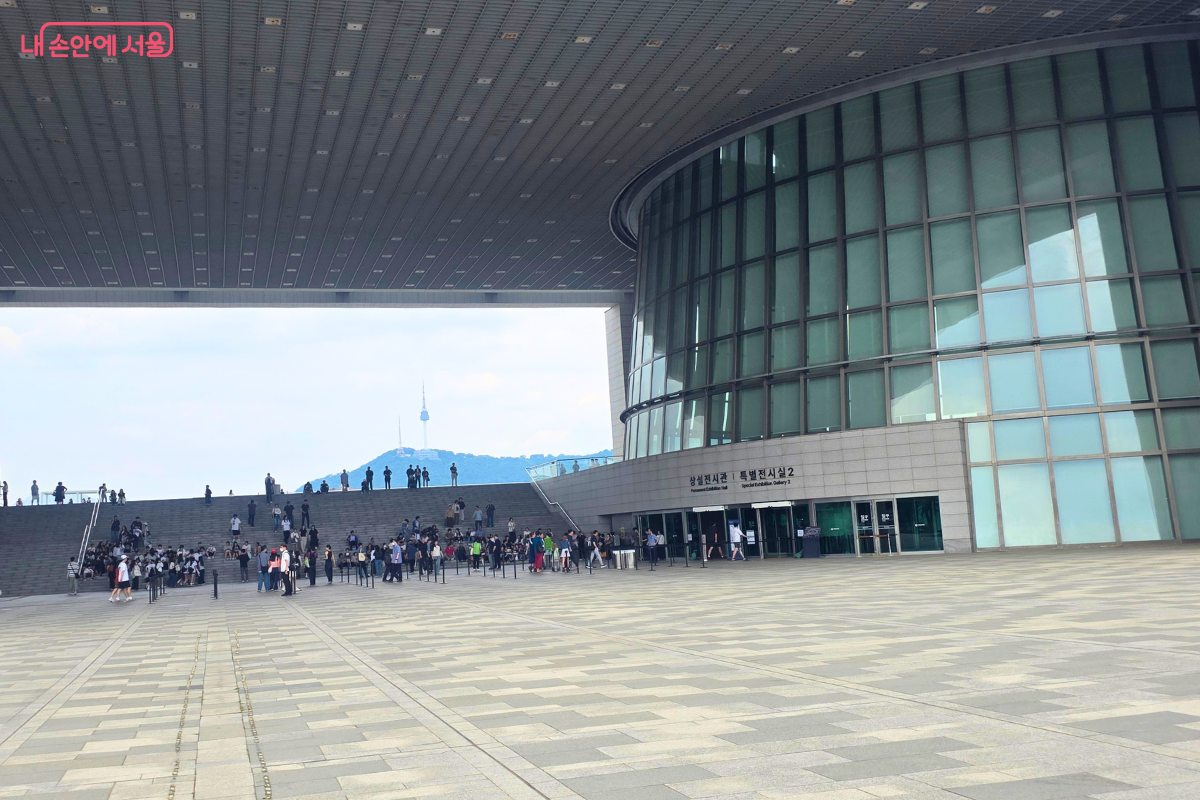 국립중앙박물관 외부 계단과 넓은 광장에 많은 시민들이 모여 있다. 박물관 건물의 큰 처마 아래로 관람객들이 이동하고, 멀리 남산과 남산타워가 보인다.