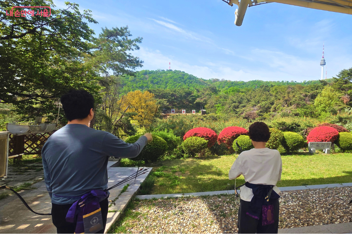 남산 석호정 사대에서 국궁 수련자들이 과녁 방향을 바라보는 모습이다. 앞쪽에는 남산의 숲이 펼쳐져 있고 멀리 N서울타워가 보인다.