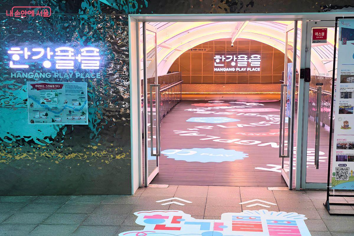 자양역 3번 출구와 연결된 한강플플 입구 전경이다. 입구 왼쪽 벽면에는 ‘한강플플 HANGANG PLAY PLACE’ 조명이 보이고, 열린 문 안쪽으로 한강플플 내부 공간과 안내 문구가 이어져 있다.