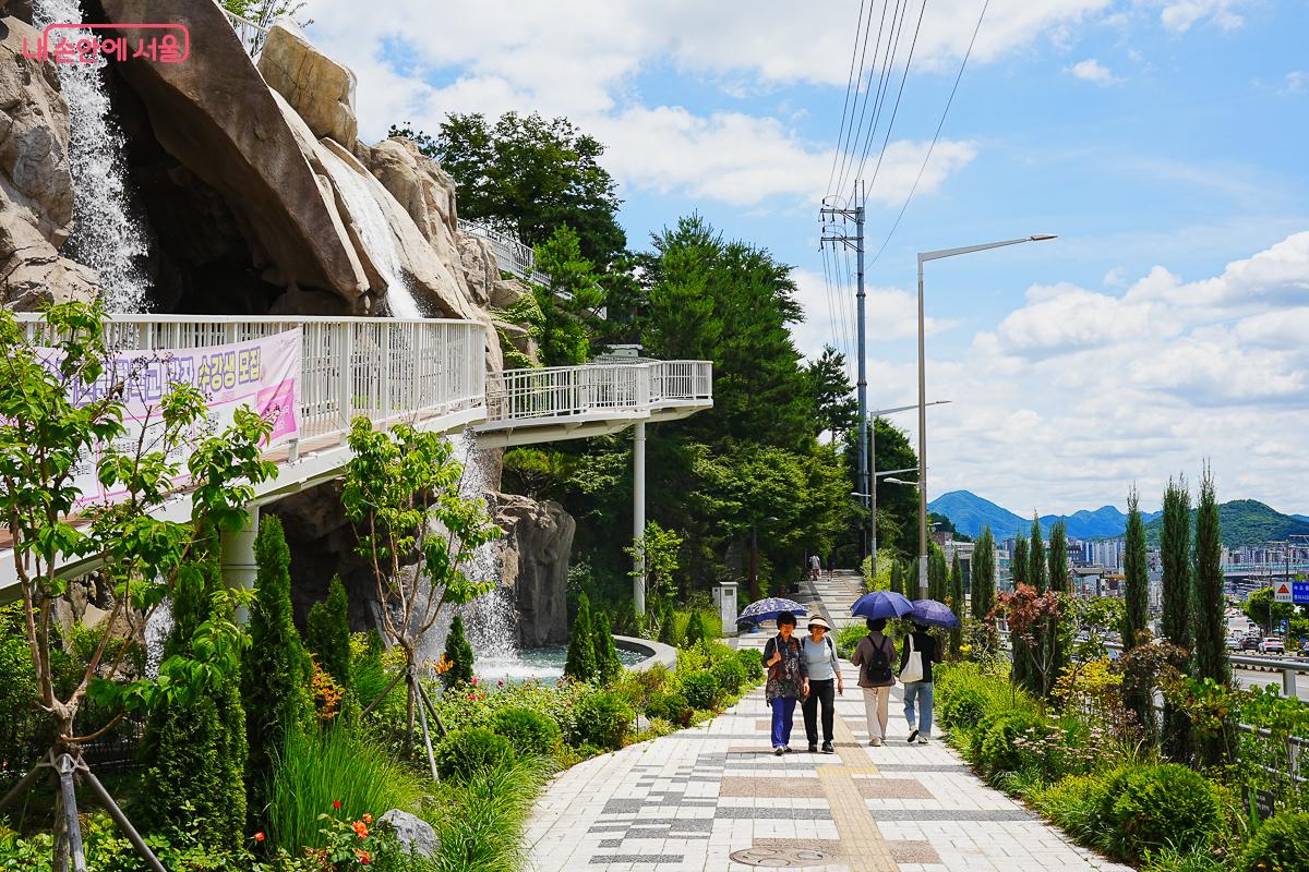 정원 산책로에서 올려다본 폭포와 나선 데크 전경, 연두빛 식물들, 배경에 관악산 능선