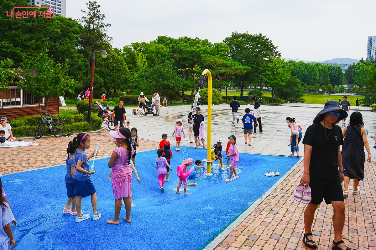 넓은 광장에 노란 샤워 기둥이 서 있고, 아이들이 오가는 여유로운 전경
