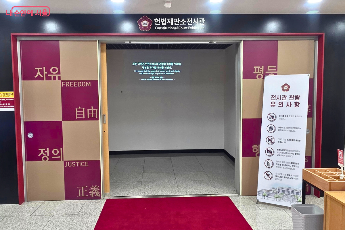 헌법재판소전시관 입구가 정면으로 보인다. 상단에는 한글과 영문 전시관 명칭이 적혀 있고, 양쪽 문에는 자유·평등·정의라는 헌법적 가치가 한글과 영문으로 표현돼 있다.