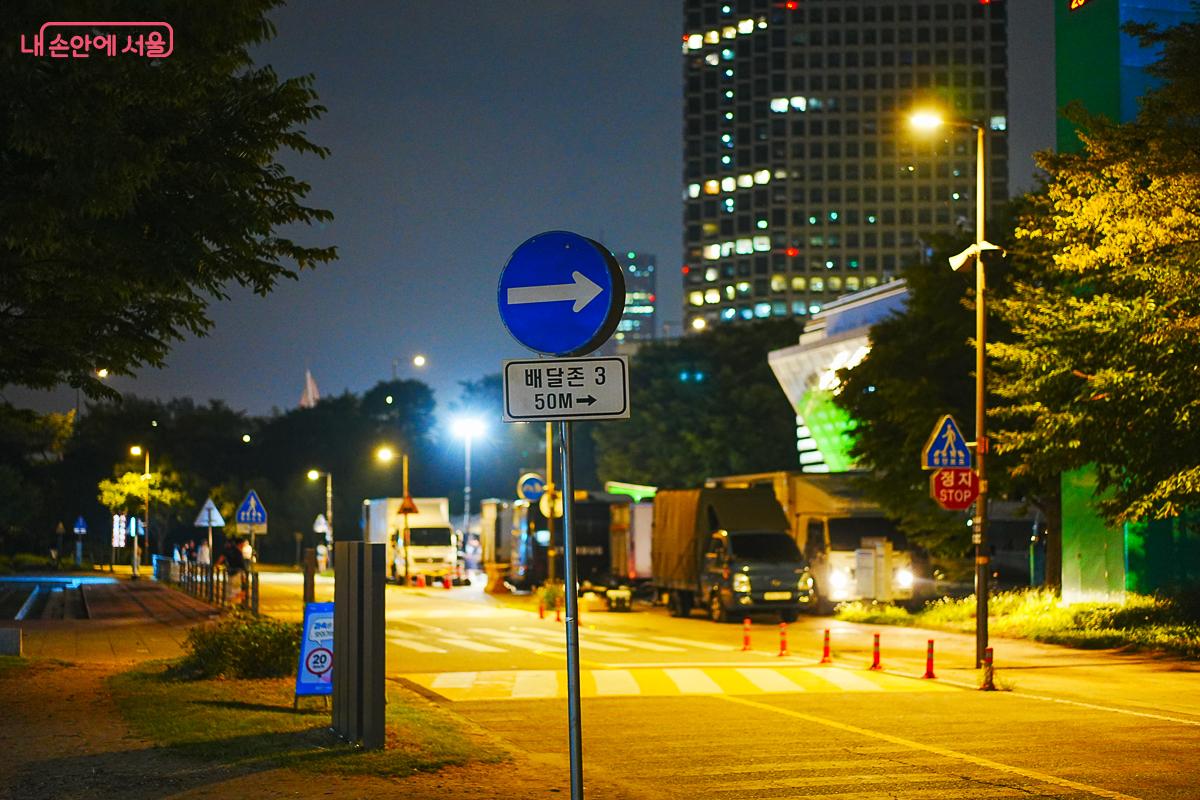 '배달존 3, 50M' 표지판과 배달 트럭들이 서 있는 야간 도로