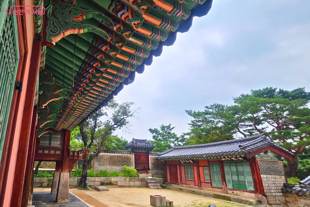 화려한 단청으로 장식된 창덕궁 전각의 처마가 왼쪽 위로 길게 이어지고, 맞은편에는 붉은 기둥과 초록색 창호를 갖춘 전각과 전통 담장이 자리한다.