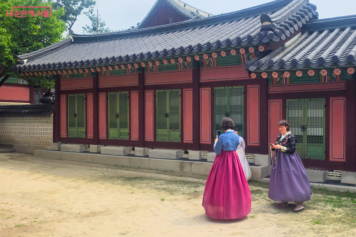 기와와 붉은 기둥, 초록색 창호가 이어진 창덕궁 전각 앞에서 화려한 색상의 한복을 입은 외국인 관람객들이 사진을 찍으며 궁궐을 둘러보고 있다.