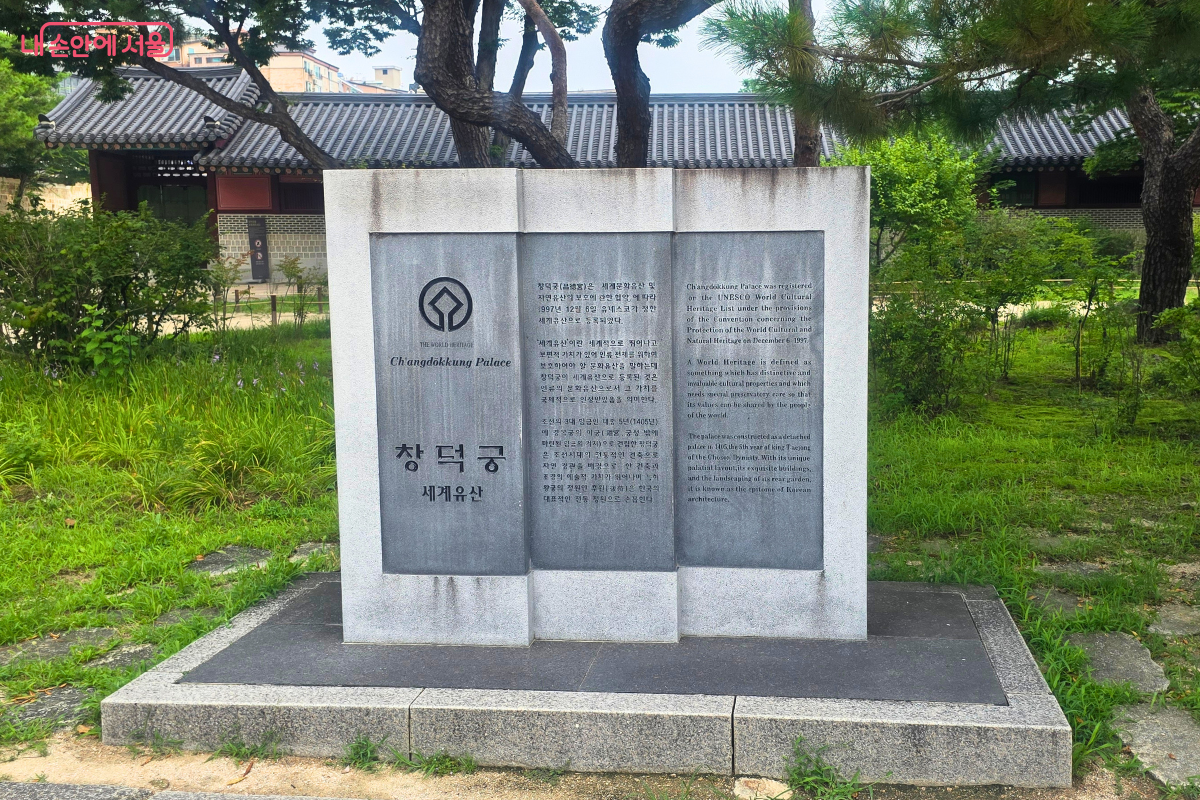 푸른 잔디와 오래된 소나무 사이에 창덕궁 세계유산 표지석이 세워져 있다. 표지석에는 유네스코 세계유산 상징과 ‘창덕궁 세계유산’이라는 글자가 새겨져 있다.