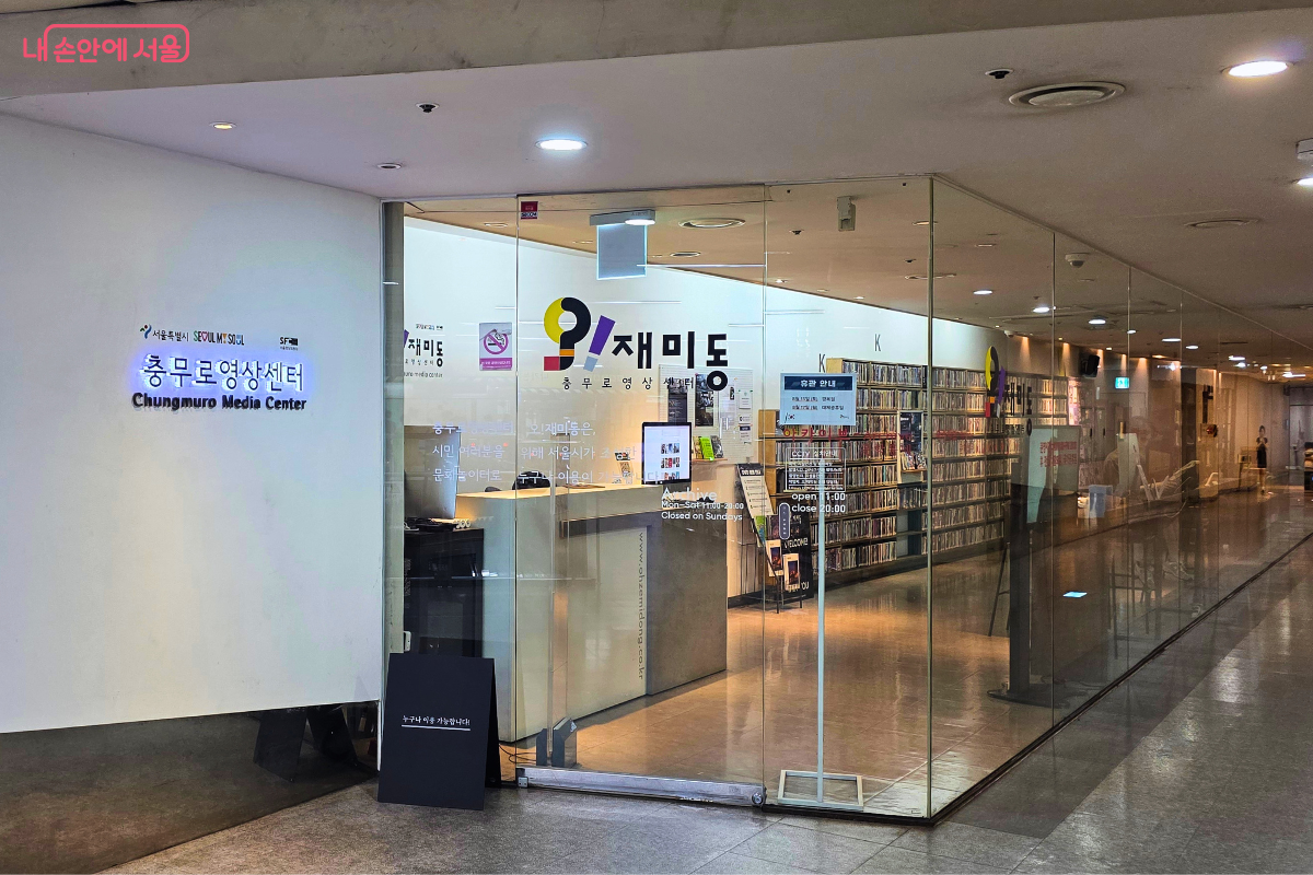 충무로역 지하 1층 통로에서 바라본 충무로영상센터 '오!재미동' 입구. 투명한 유리벽과 출입문 너머로 안내 데스크와 DVD 서가가 보인다.