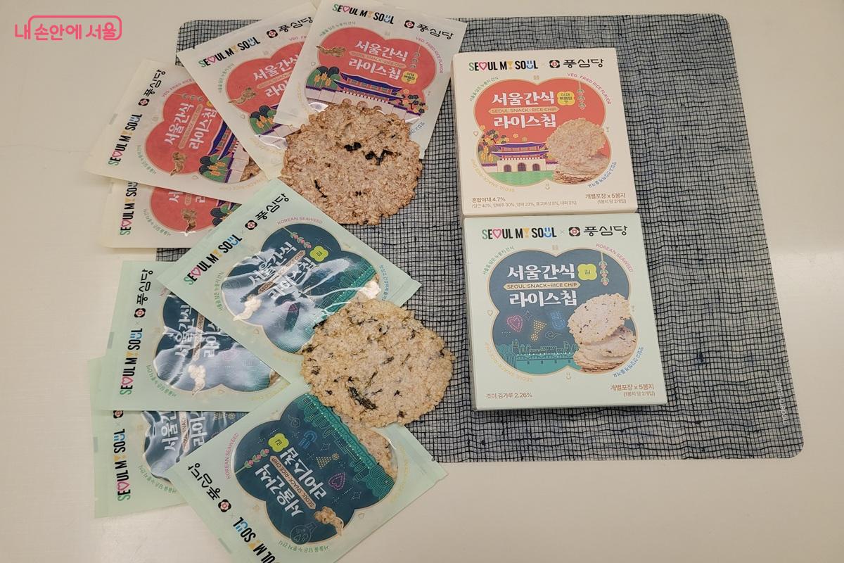 야채볶음밥 맛과 김 맛 라이스칩의 상자, 개별 포장지, 둥근 과자가 식탁에 놓여 있다.