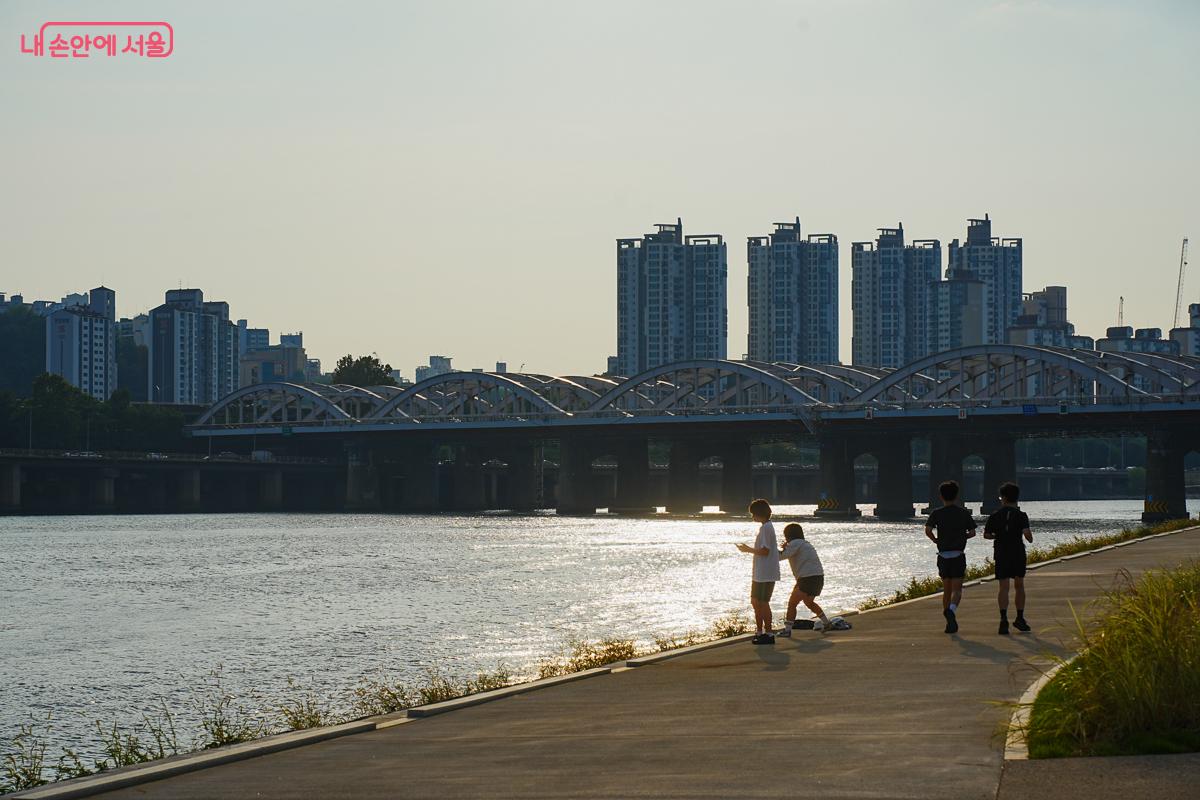 노을을 등지고 한강변을 달리는 러너들과 산책하는 사람들의 실루엣, 한강철교가 보인다.