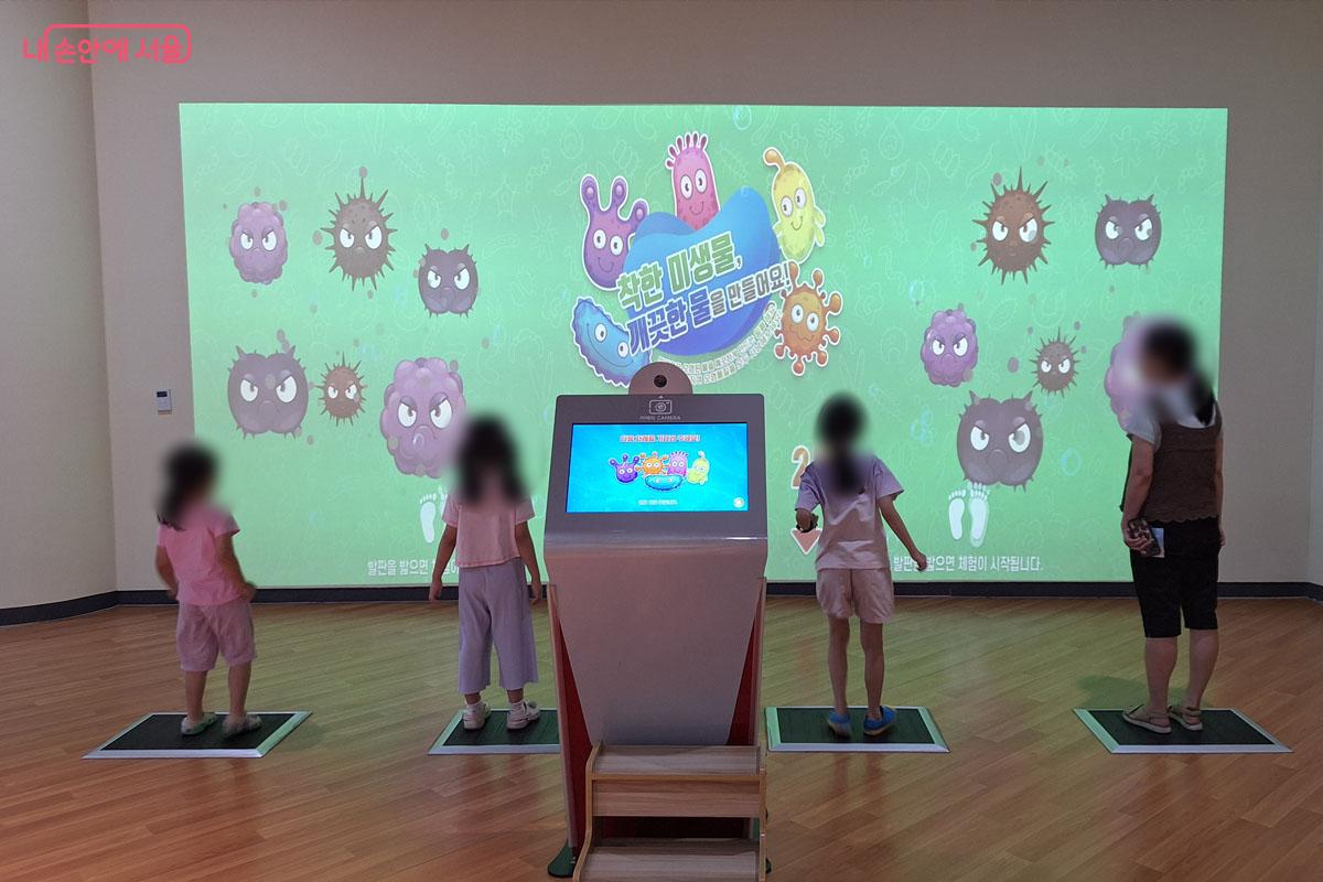 아이들이 발판 위에 서 있고 벽 스크린에는 화면이 나오고 있다. 나쁜 세균들을 물리치는 것 같은 그림이 그려져 있다.