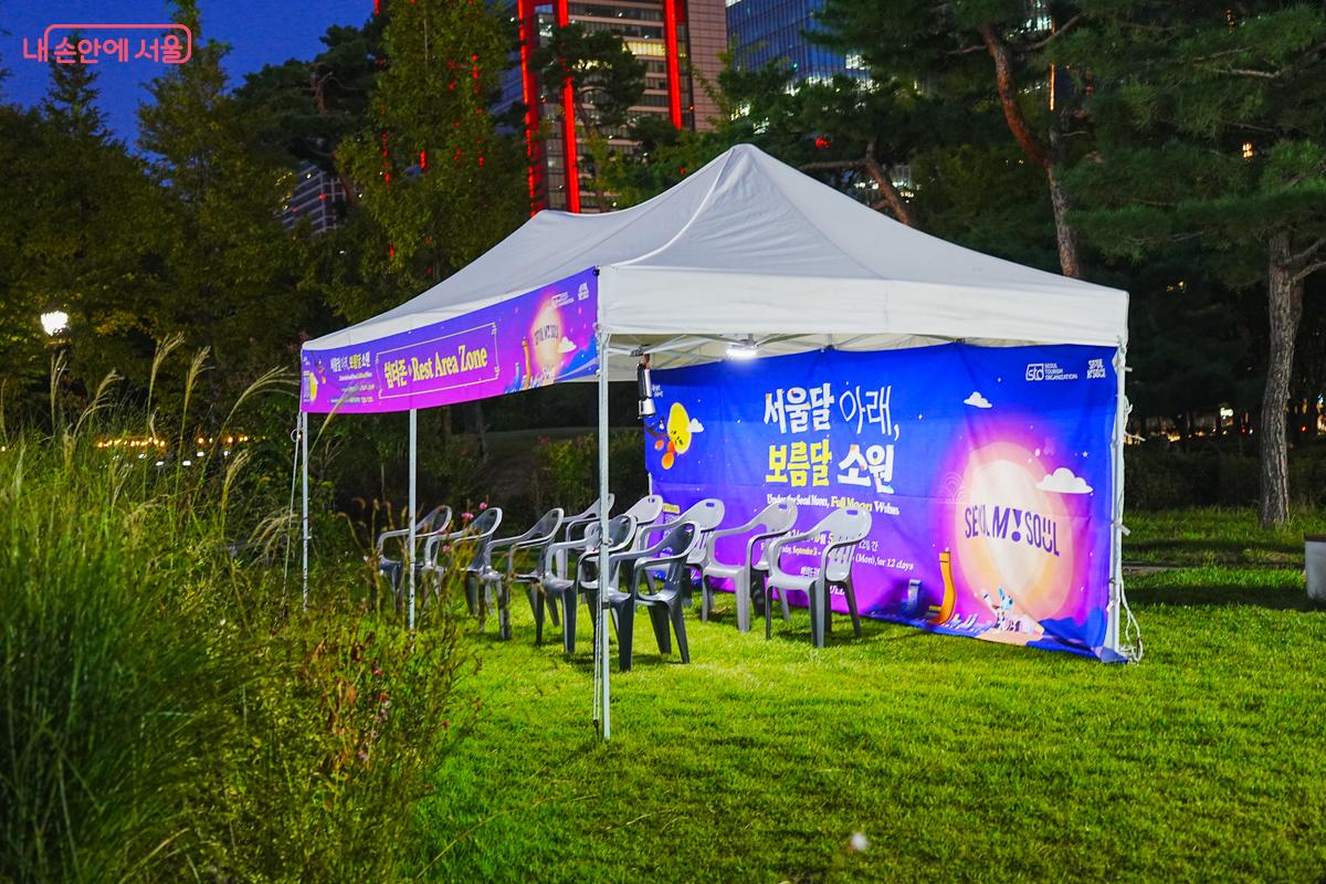 '휴식존 Rest Area Zone' 배너가 걸린 하얀 텐트와 잔디밭에 놓인 의자들, 뒤로 도심 빌딩이 보인다.