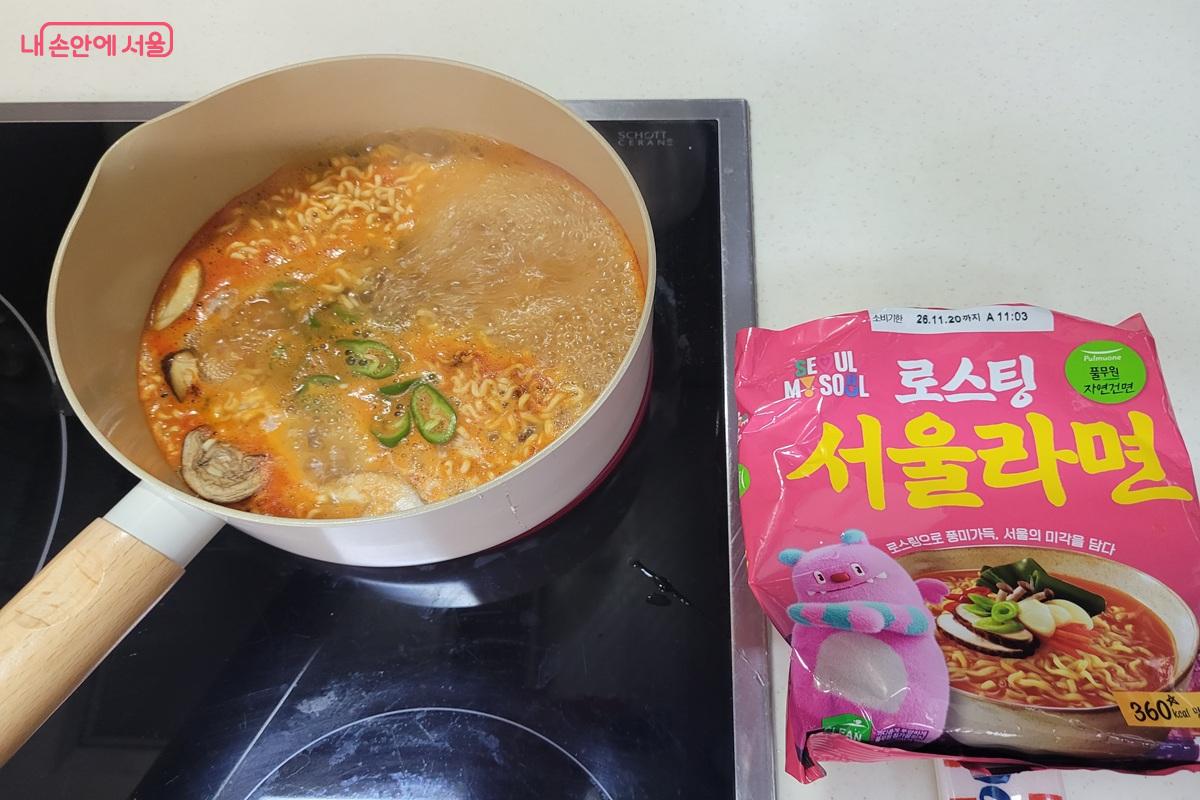 냄비에서 서울라면이 버섯과 채소와 함께 끓고 있고 옆에 분홍색 서울라면 봉지가 놓여 있다.