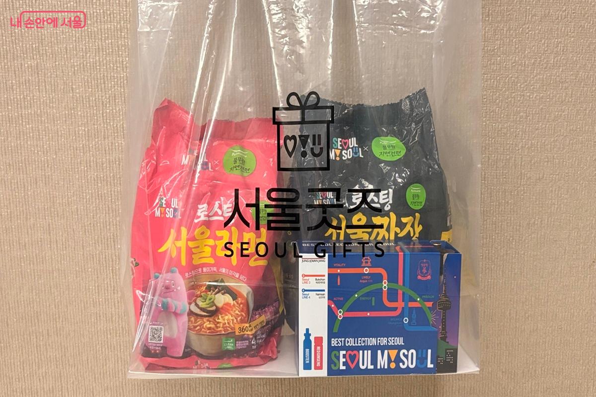 투명한 서울 굿즈 쇼핑백 안에 분홍색 서울라면, 검은색 서울짜장, 지하철 노선도 디자인의 홍삼음료 상자가 담겨 있다.