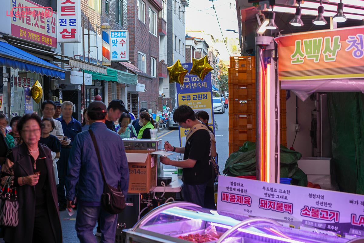금색 별 풍선이 달린 '주류 교환처' 간판 아래 사람들이 줄을 서 있고, 옆으로 정육점의 화려한 메뉴 네온사인이 보인다.