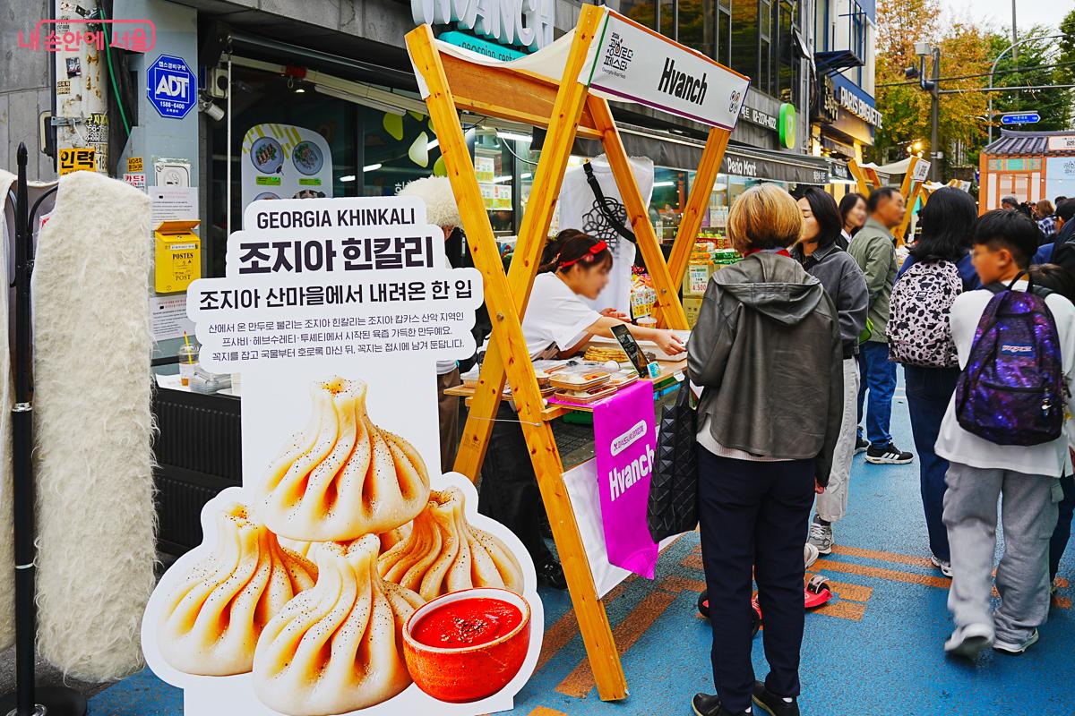 GEORGIA KHINKALI 조지아 흔칼리' 안내판과 만두 사진이 세워진 부스, 상인이 음식을 준비하고 있다.