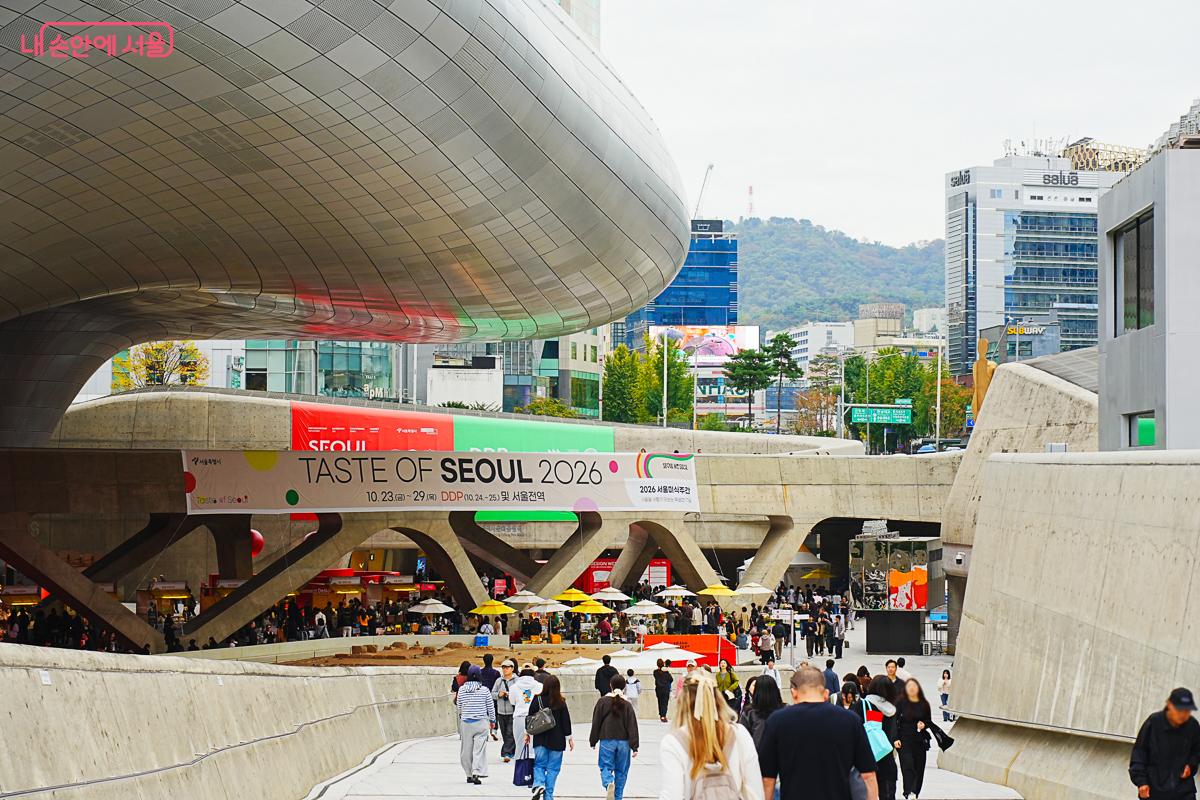 동대문디자인플라자(DDP)의 곡선형 외관 아래로 'TASTE OF SEOUL 2026' 현수막이 걸려 있고, 그 아래로 노란 파라솔이 늘어선 장터와 시민들이 보인다.