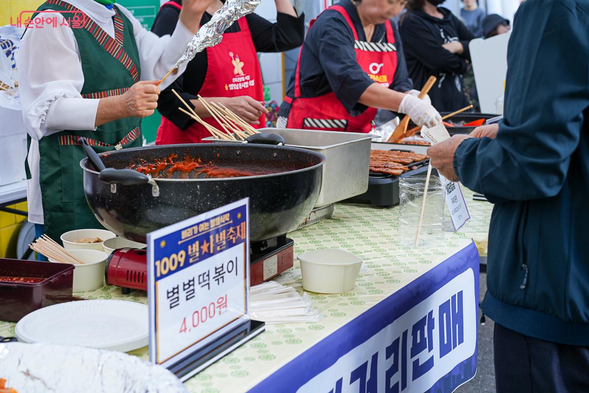 '1009 별★별축제' 로고와 '별별떡볶이 4,000원' 가격표가 놓인 좌판, 상인들이 큰 팬에 떡볶이를 볶는 모습