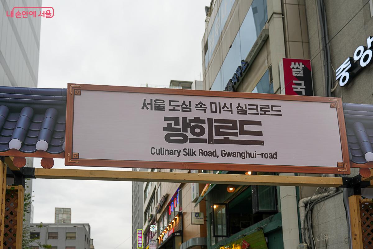 기와지붕 모양의 아치형 구조물에 걸린 안내판, '서울 도심 속 미식 실크로드 광희로드 / Culinary Silk Road, Gwanghui-road' 문구가 적혀 있다.