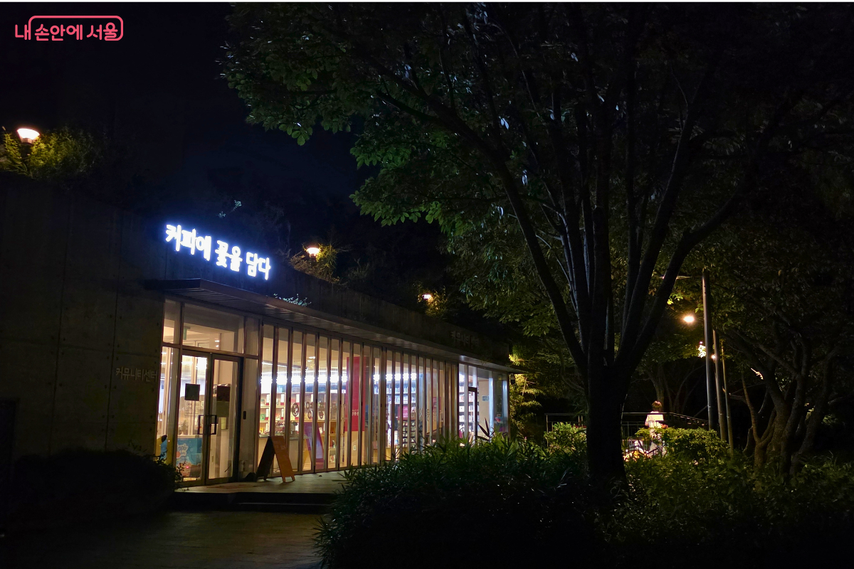 어두운 서울숲 숲길 옆으로 &lsquo;커피에 꽃을 담다&rsquo;라는 밝은 간판이 보이는 유리 외벽 건물이 있다.
