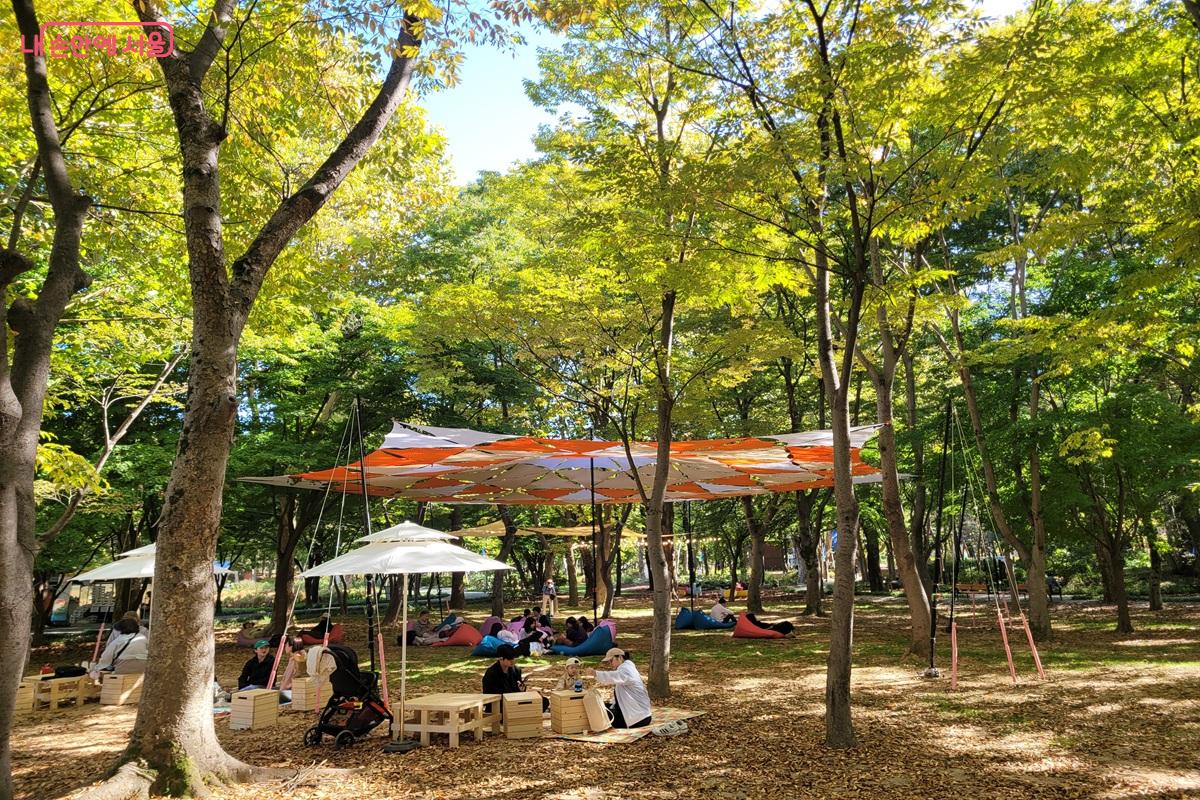 매헌시민의숲의 주황색과 흰색 차양 아래 빈백에 기대거나 파라솔 아래 탁자 주변에 앉아 쉬는 시민들