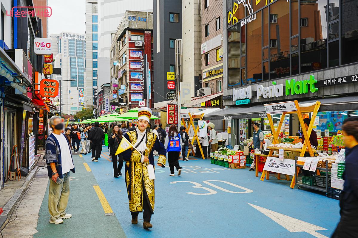 금빛 전통 의상을 입고 깃발을 든 남성이 거리를 걷고, 오른쪽으로 과일&middot;채소 노점과 'KHINKALI' 간판이 보인다.