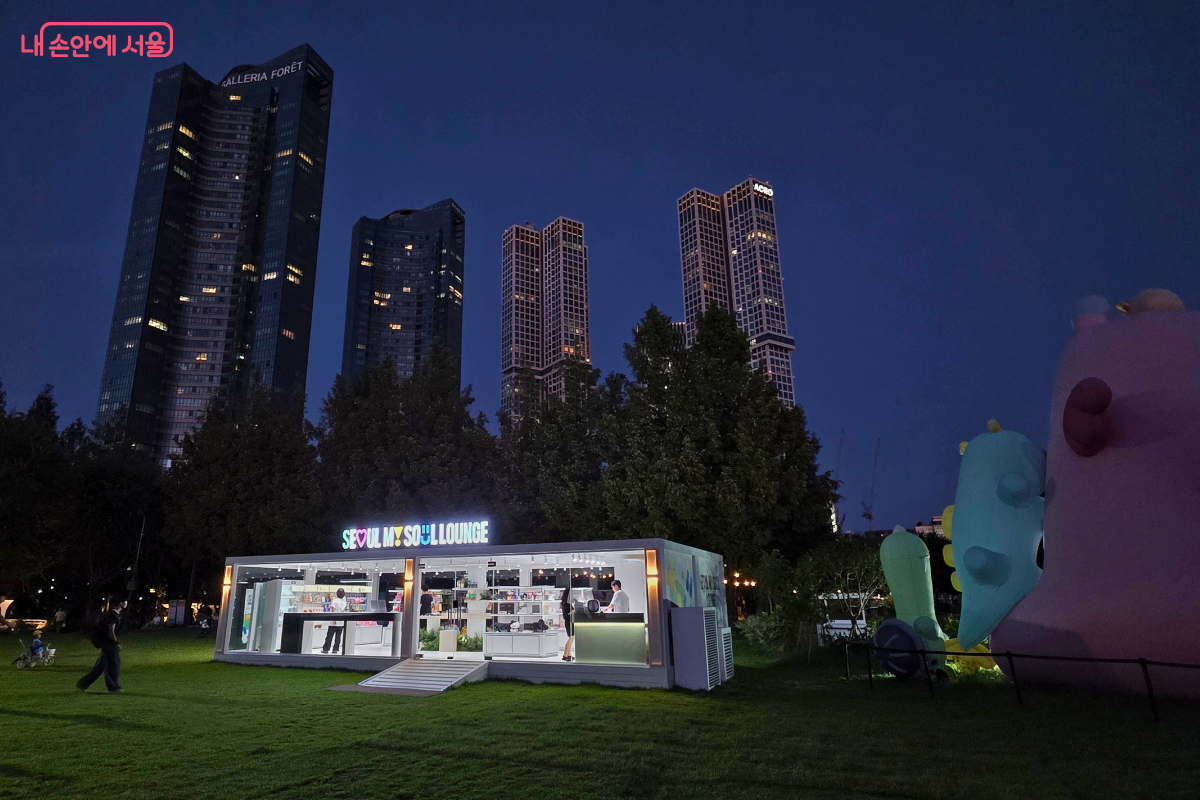 밤하늘 아래 서울숲 잔디광장에 &lsquo;SEOUL MY SOUL LOUNGE&rsquo; 건물이 환하게 빛나고 있다. 뒤로는 고층 빌딩들이 서 있고, 오른쪽에는 대형 설치물이 보여 도심 속 공원의 야경이 돋보인다.