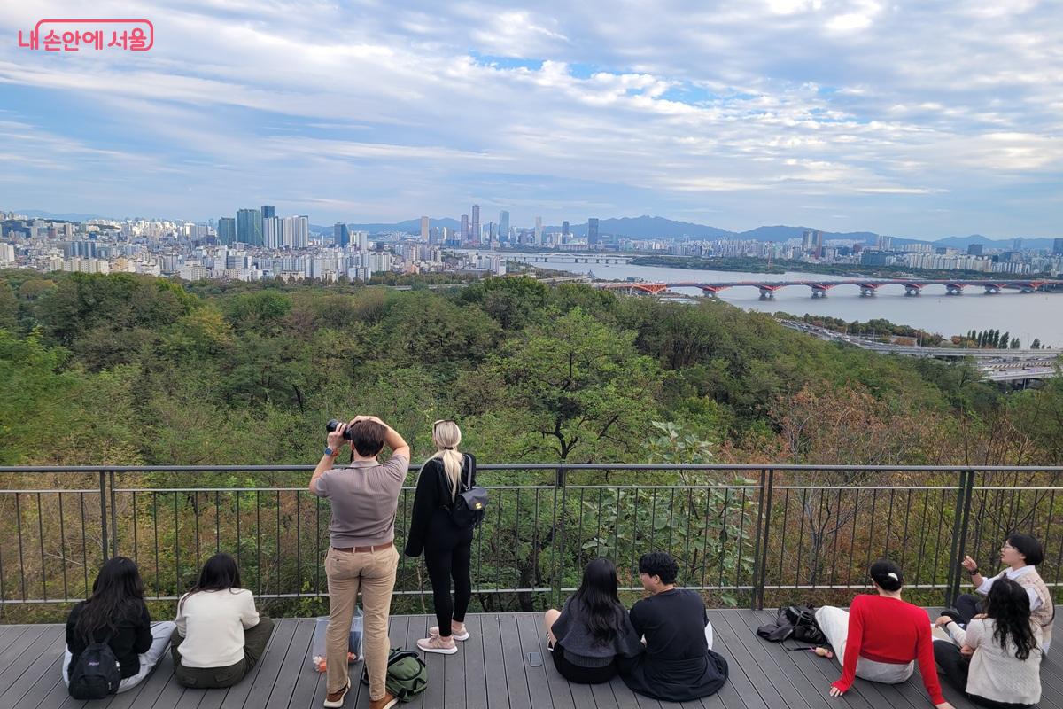 전망대에 앉거나 서 있는 시민들 앞쪽으로 한강과 다리, 서울의 건물들이 펼쳐져 있다.